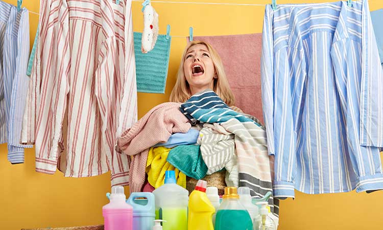 Waschmittelpulverflecken entfernen, Frau verzweifelt an großem Wäscheberg mit vielen Waschmittelflaschen